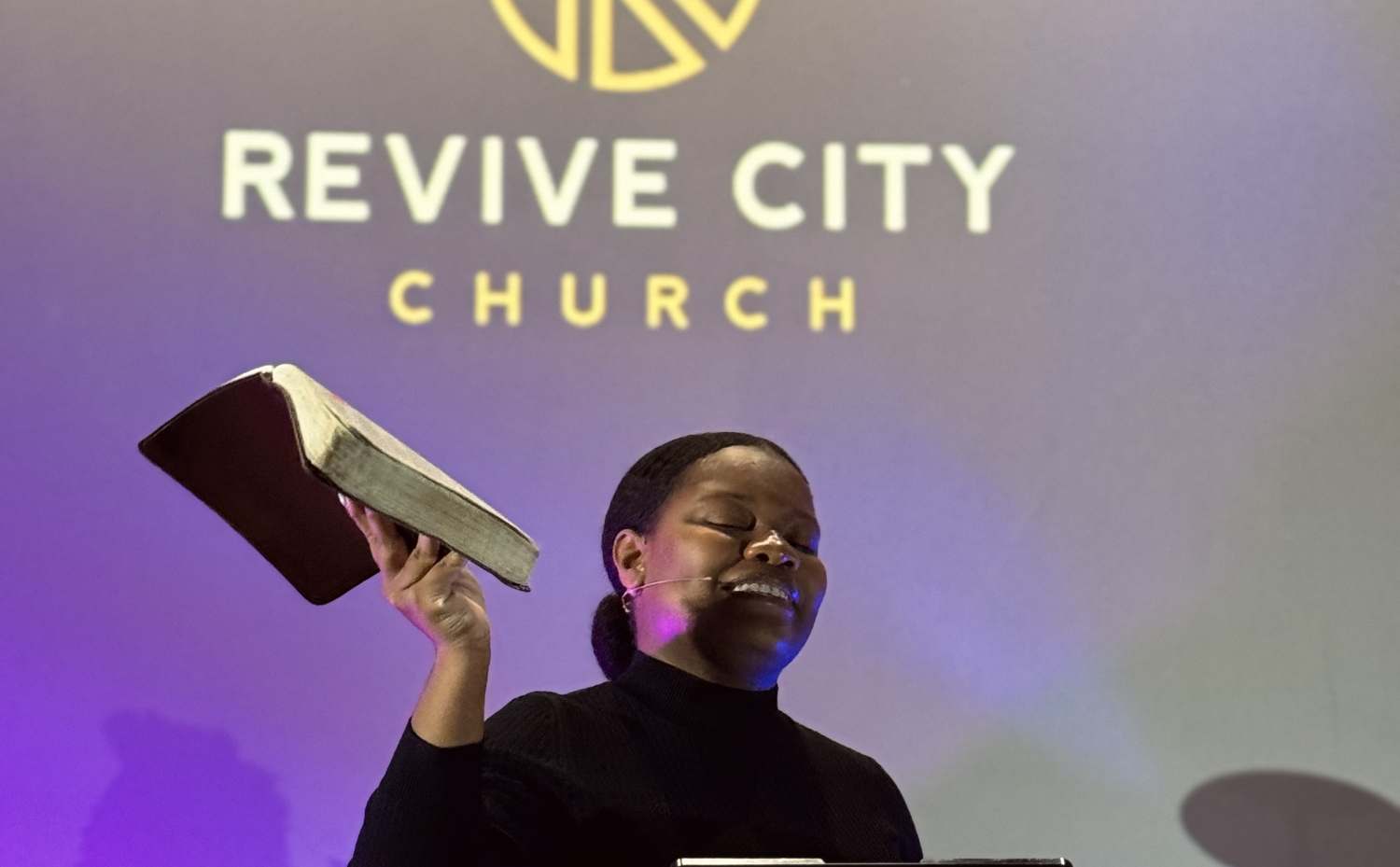 Pastor Rebecca Jean preaching with her Bible raised at Revive City Church
